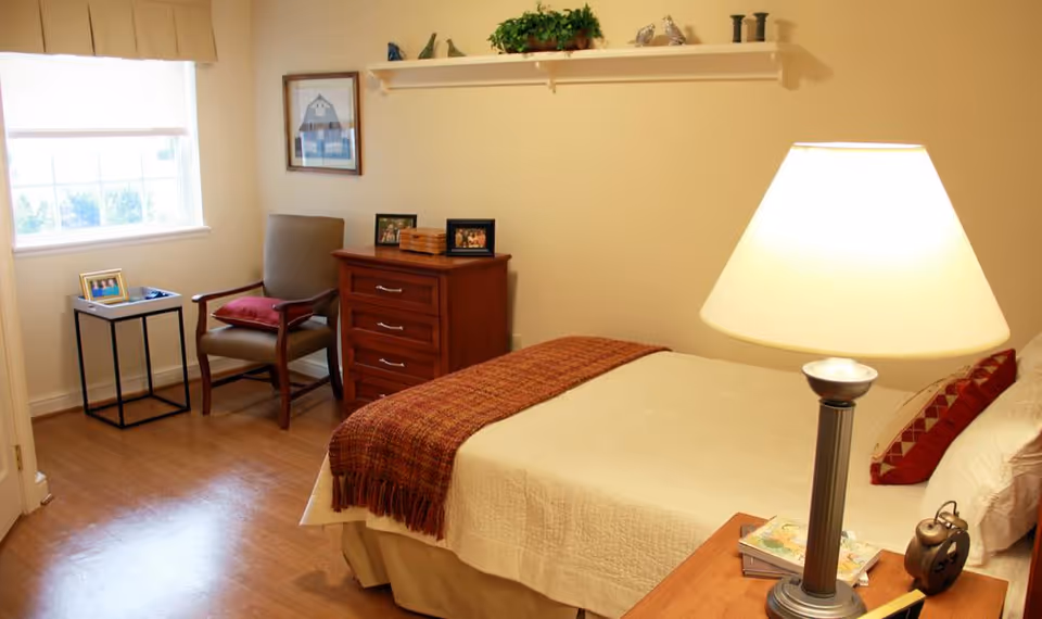 A cozy bedroom in a senior living facility featuring a bed with a beige quilt and a red throw blanket, a bedside table with a lamp and an alarm clock, a wooden dresser with framed photos, a cushioned chair with a red pillow, a small side table with a framed photo, and a window with a beige valance letting in natural light.