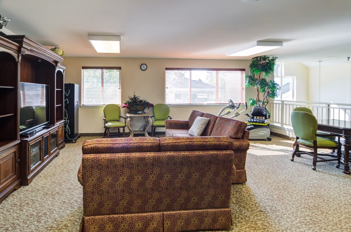 A bright senior living common room with patterned sofas facing a wooden entertainment center, green chairs by the windows, a table, and an exercise machine.
