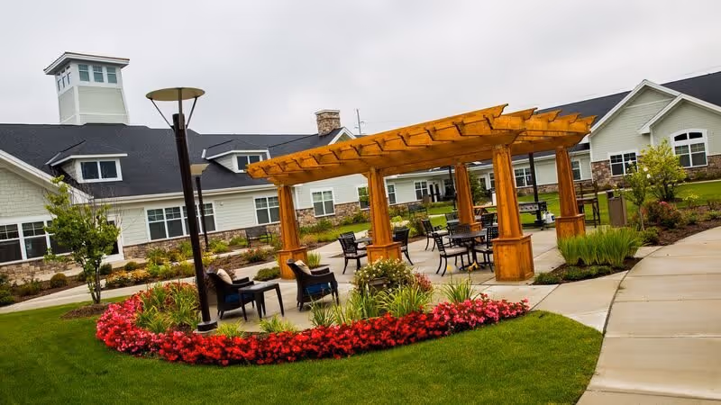 Outdoor patio area at a senior living facility featuring a wooden pergola with tables and chairs underneath, surrounded by well-maintained green lawns, colorful flower beds, and a paved walkway. The background shows a large building with white siding and stone accents.