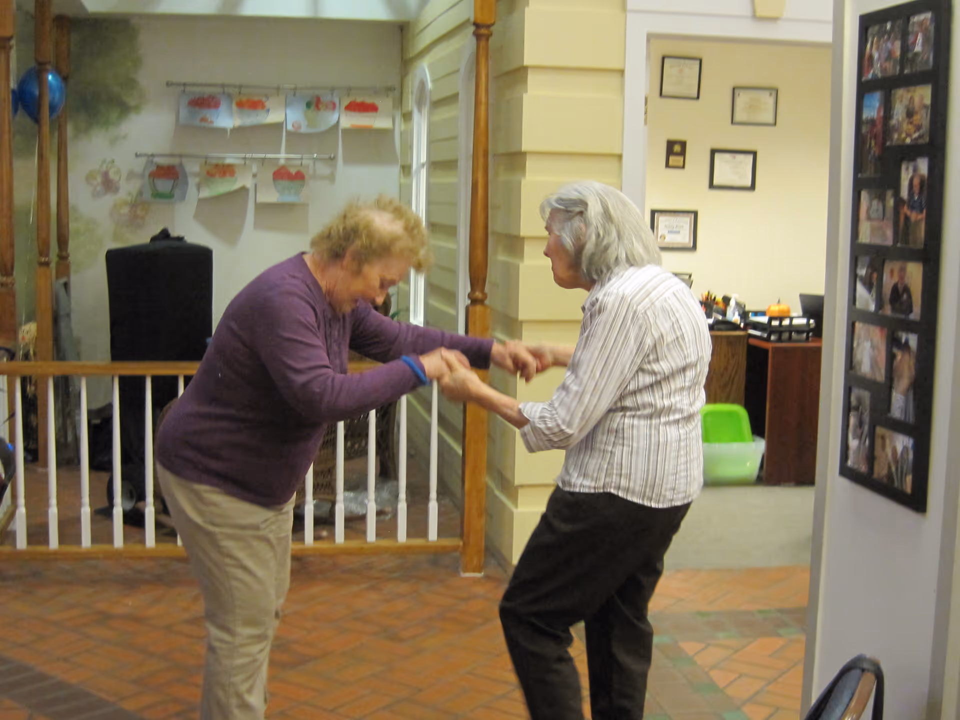Two elderly women holding hands and dancing together indoors in a senior living facility. The room has a brick floor, a white railing, and walls decorated with children's artwork and framed certificates.
