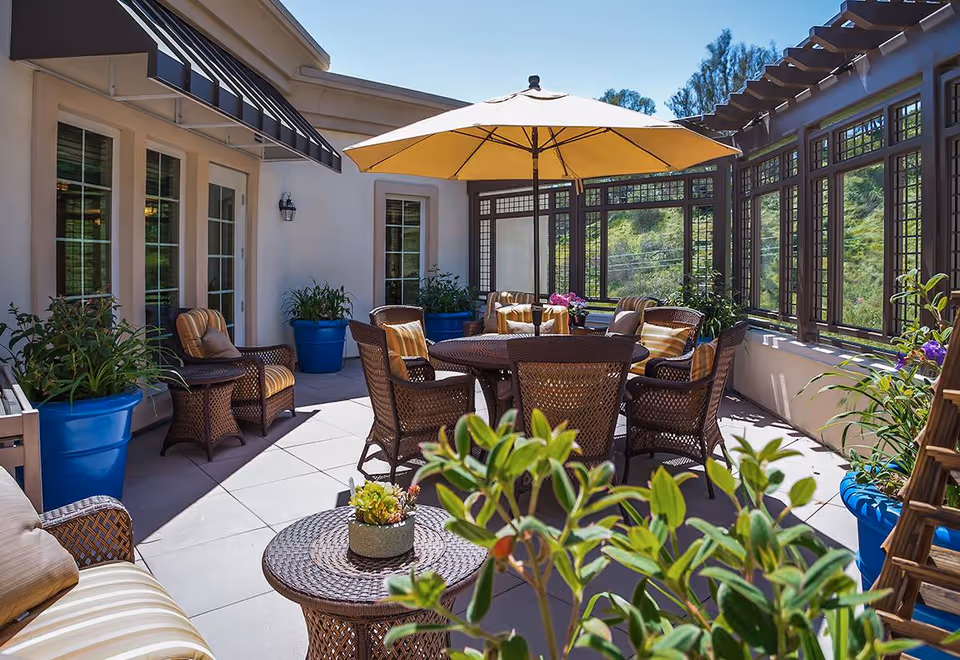 Sunlit outdoor patio with wicker chairs and a round table under a large yellow umbrella, surrounded by potted plants and windows.