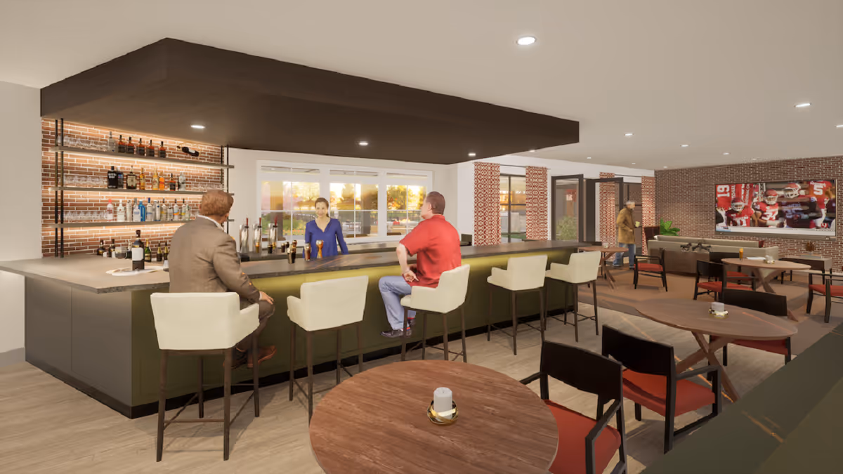 Interior view of a senior living facility lounge area with a bar counter where two men are seated and a bartender is standing behind the bar. The room has several tables and chairs, a large TV on the brick wall showing a football game, and a man walking in the background.