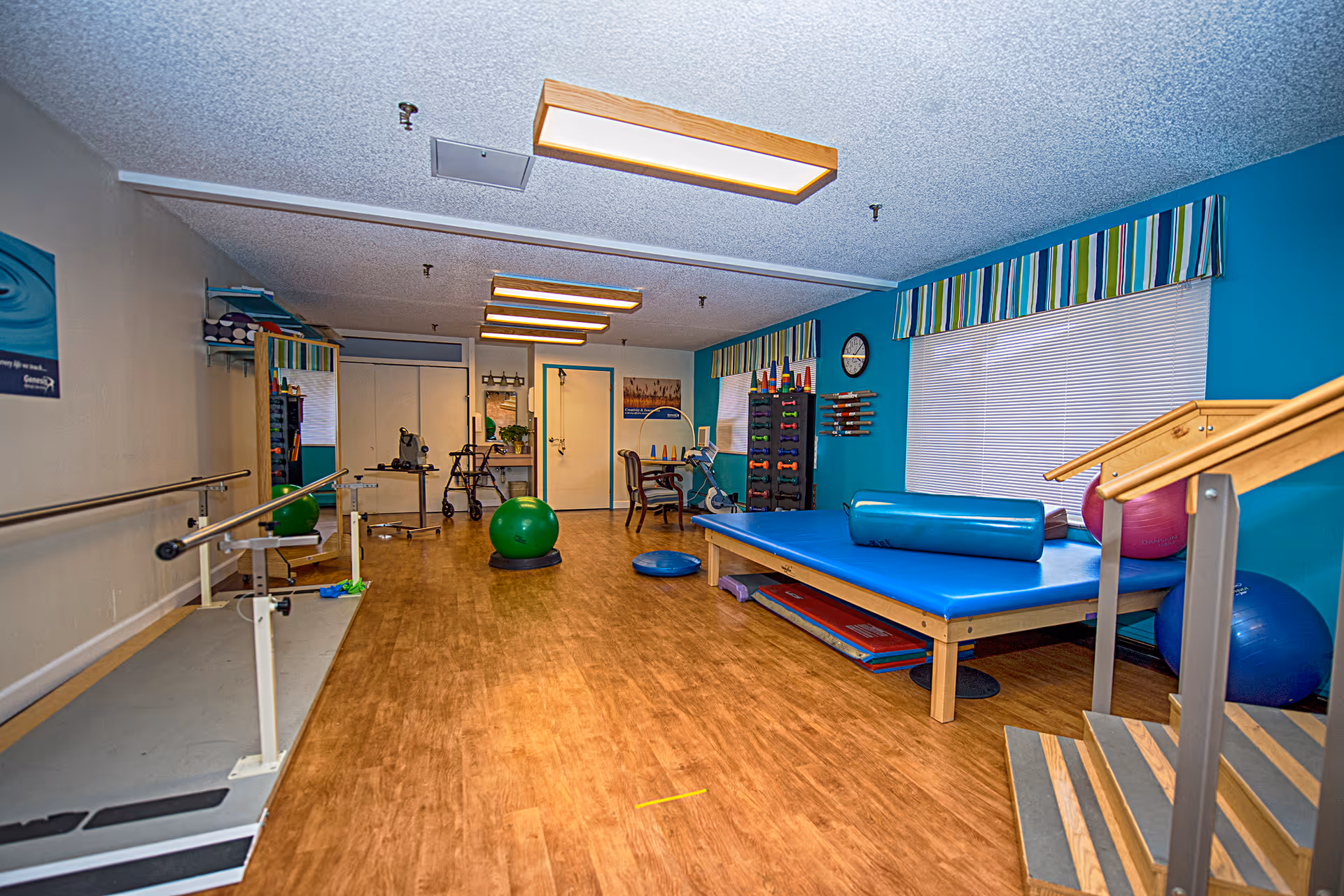 A rehabilitation therapy room with wooden flooring and blue walls. The room contains exercise equipment including parallel bars, a large blue therapy mat with cylindrical cushions, various exercise balls, hand weights on a rack, and a set of stairs for physical therapy. There is a window with striped valances and a clock on the wall. The room is well-lit with ceiling lights.
