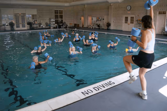 A group of elderly individuals participating in a water exercise class in an indoor swimming pool, following the instructor who is standing poolside holding blue foam dumbbells.