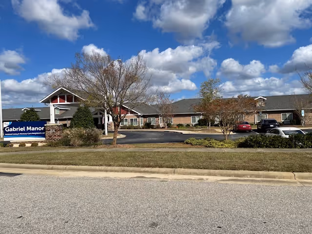 Exterior view of Gabriel Manor Assisted Living Center, a single-story brick building with a pitched roof under a partly cloudy blue sky. There is a parking lot with several cars and a sign in front that reads 'Gabriel Manor Assisted Living & Memory Care Neighborhood'. Trees and shrubs are planted around the building and parking area.