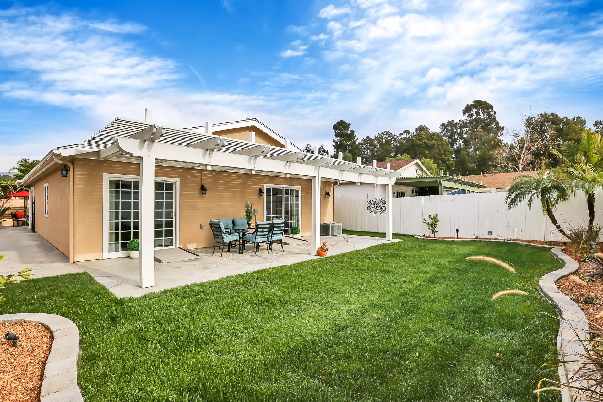 Backyard of a senior living facility with a well-maintained green lawn, a white pergola covering a patio area with outdoor dining furniture, beige exterior walls, sliding glass doors, and a white fence with some plants and trees around the perimeter under a partly cloudy blue sky.