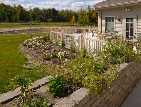 A fenced patio and seating area attached to a beige single-story building beside a landscaped flower bed and grassy field.