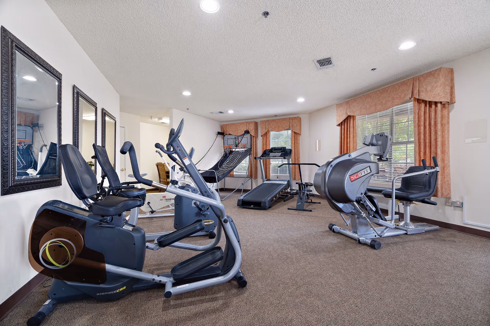 A fitness room in a senior living facility with exercise equipment including stationary bikes, a treadmill, and a rowing machine. The room has beige carpet, white walls, three framed mirrors on one wall, and windows with peach-colored curtains allowing natural light to enter.