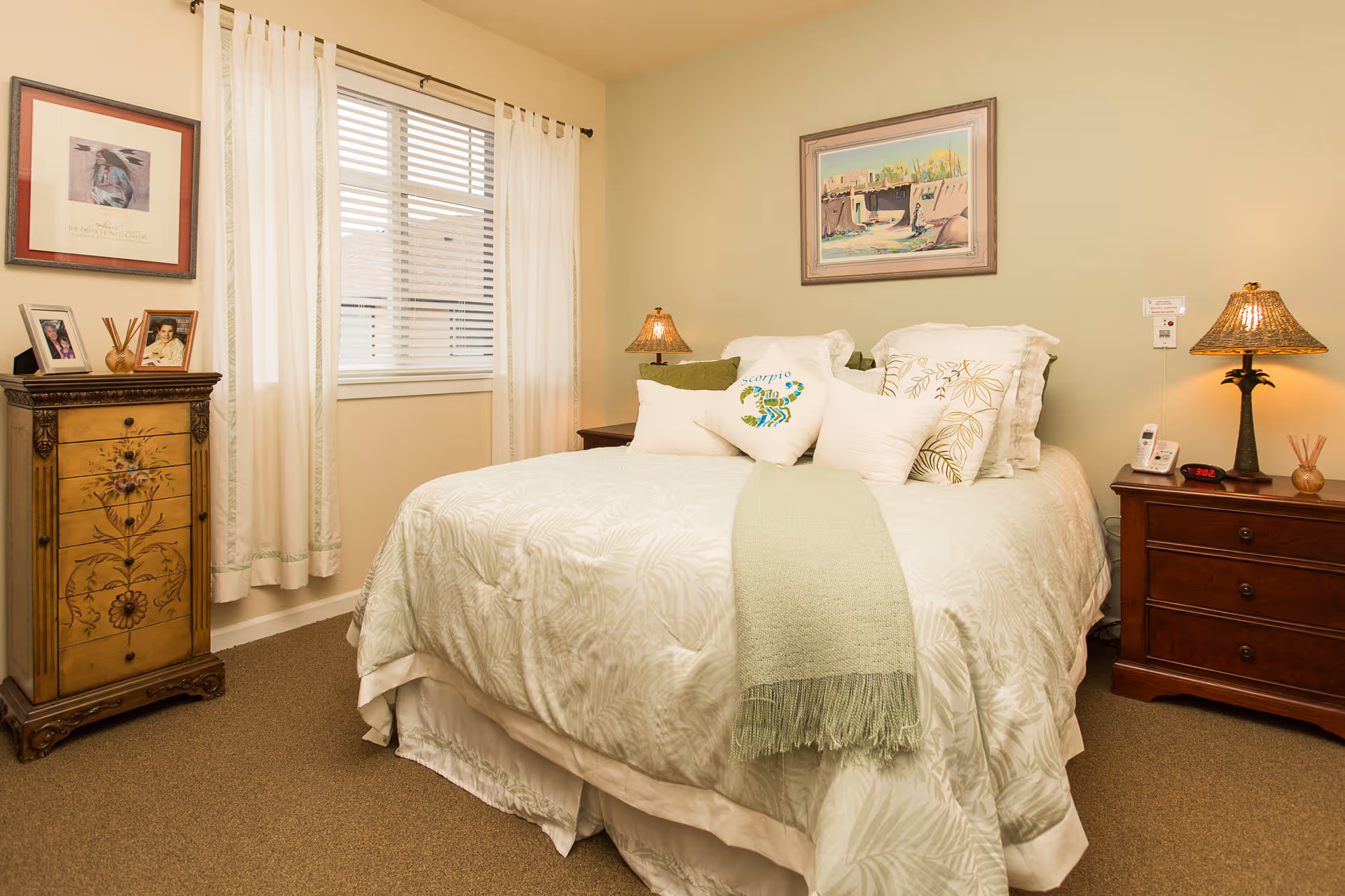 A cozy bedroom with a neatly made bed featuring white and light green bedding and multiple pillows, including one with a scorpion design. There are two wooden nightstands on either side of the bed, each with a lamp. A window with white curtains is on the left wall, and framed artwork hangs above the bed and on the adjacent wall. A decorative wooden chest with framed photos and a reed diffuser is positioned near the window.