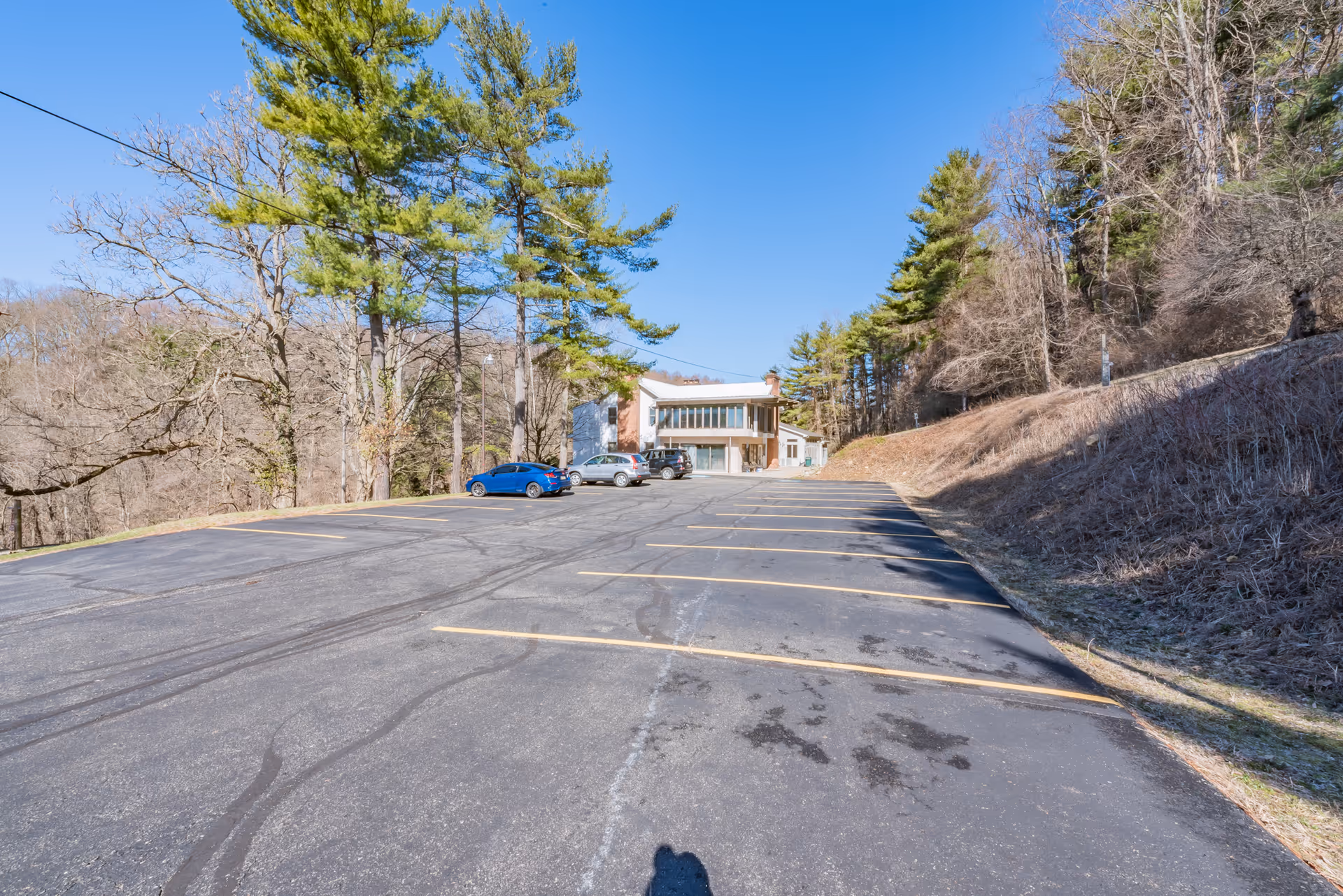 A large parking lot with a few parked cars in front of a two-story building surrounded by trees under a clear blue sky.