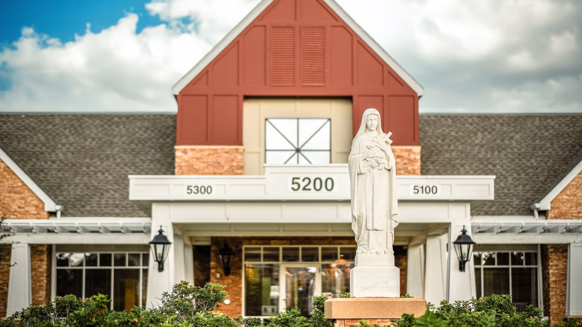 Front exterior view of Saint Therese Senior Living of Oxbow Lake building featuring a statue of a saint holding a cross in the foreground, with a brick and red panel facade, large windows, and the address numbers 5100, 5200, and 5300 displayed above the entrance under a partly cloudy sky.
