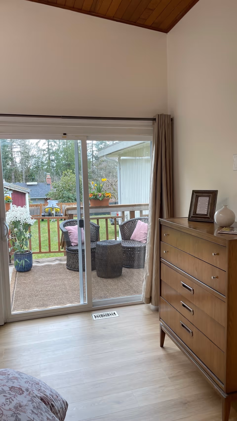 View of a room with a wooden dresser on the right and a sliding glass door leading to an outdoor patio with two wicker chairs, a small table, and potted plants. The patio overlooks a grassy backyard with trees and neighboring houses.