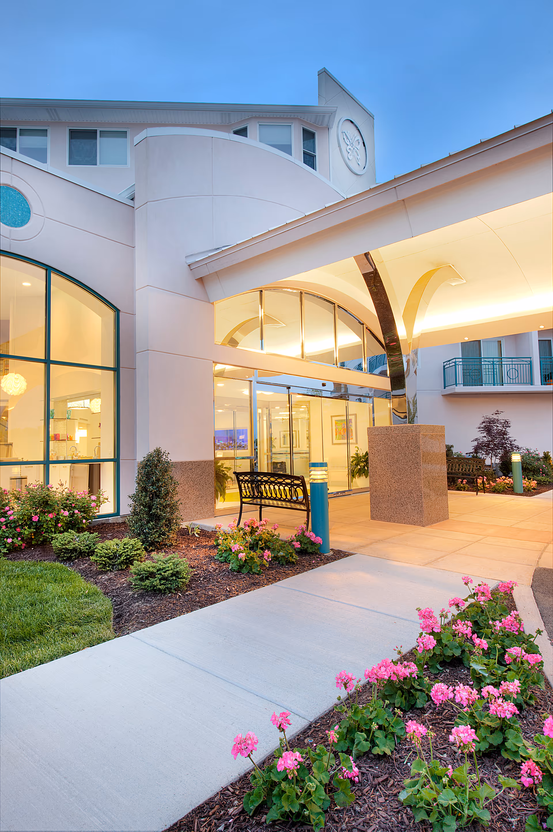 Entrance of a senior living building with a covered glass doorway, a bench, landscaped flower beds and illuminated canopy at dusk.