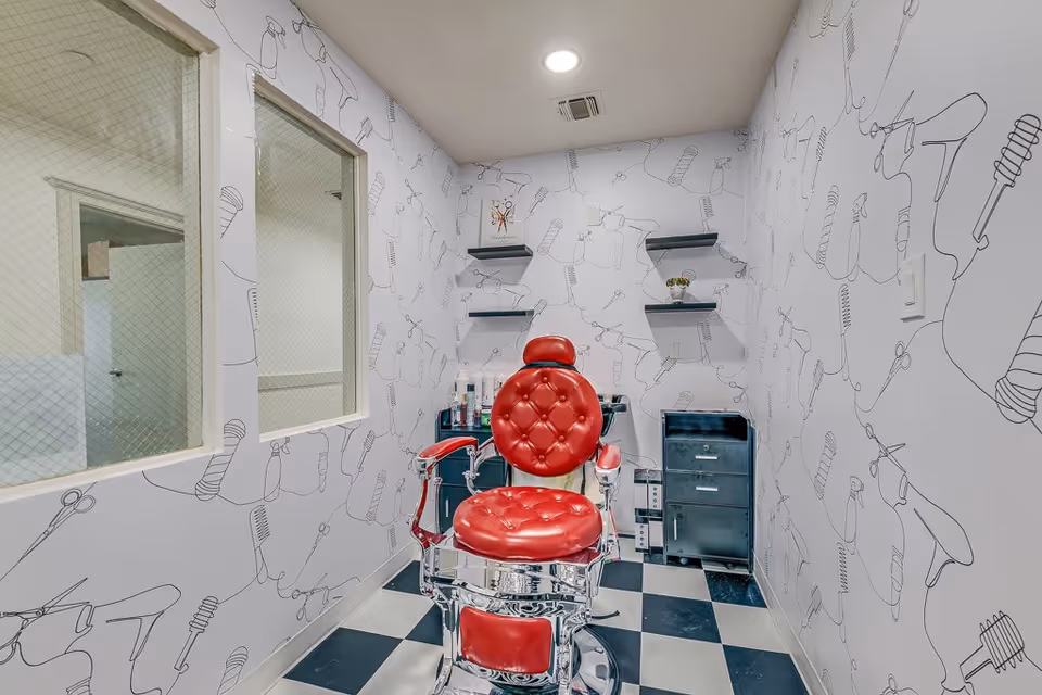 A small salon room with a vintage red barber chair in the center. The walls are decorated with black and white illustrations of hair styling tools like scissors, combs, and hair dryers. There are three black shelves on the back wall holding small items, and a black cabinet with drawers is positioned beneath them. The floor has a black and white checkered pattern, and two windows with wire mesh are on the left wall.