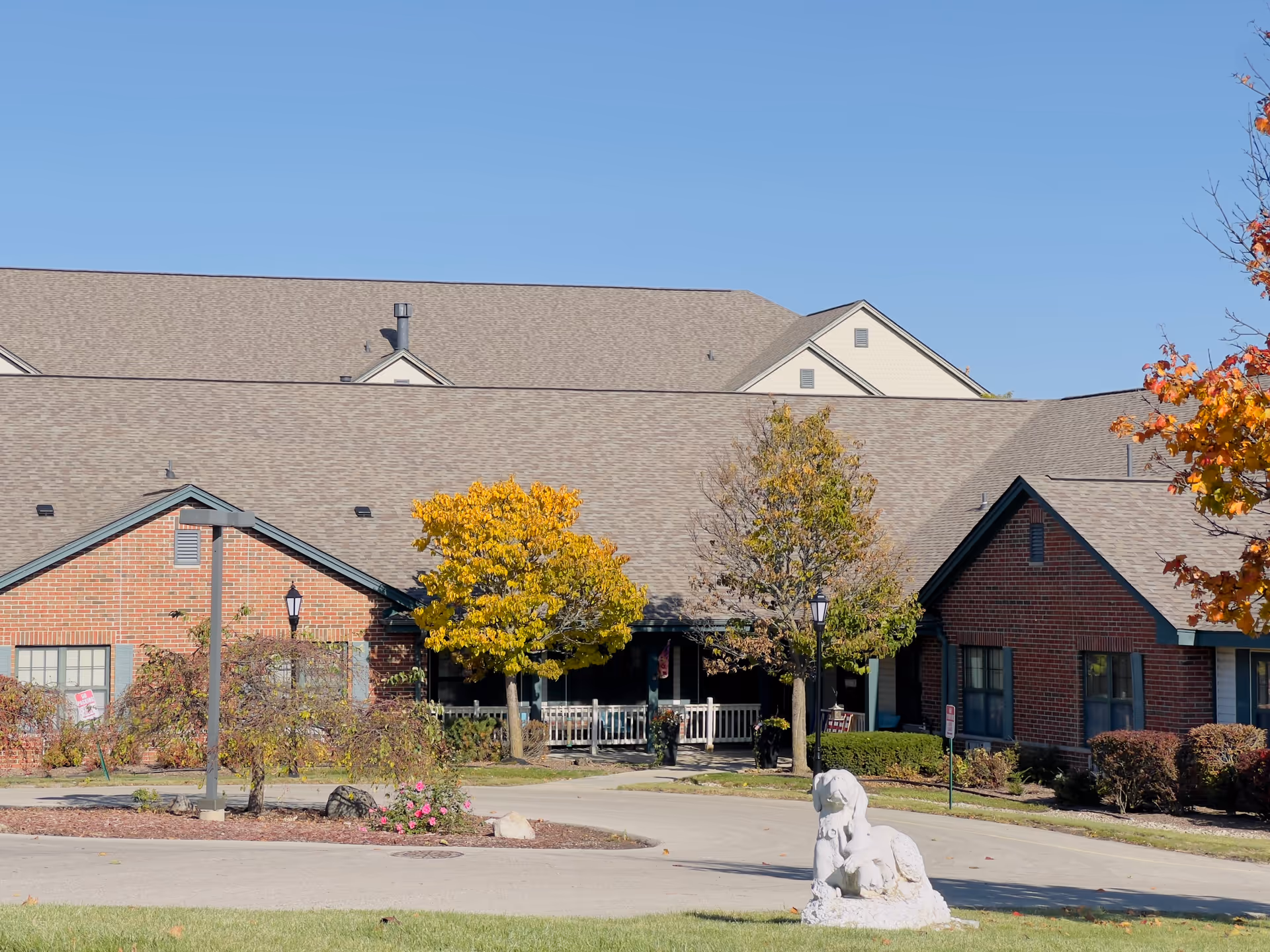 Front exterior of a brick senior living building with trees in fall colors, a porch entrance, and a small stone dog statue on the lawn.