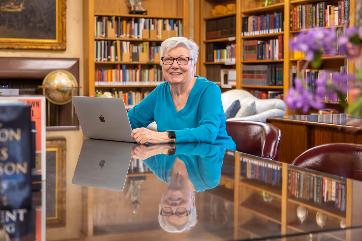 An elderly woman with short white hair and glasses sits at a polished table with a laptop in front of her, smiling. Behind her are bookshelves filled with books and a globe, suggesting a library or study room setting. Purple flowers are visible in the foreground on the right side.