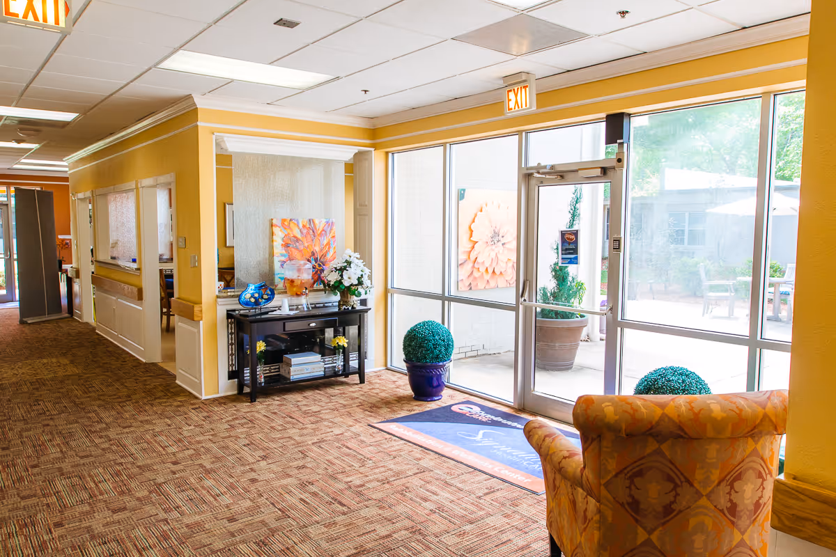 Bright senior living facility lobby with an upholstered chair, console table with flowers and decor, and glass doors leading outside.