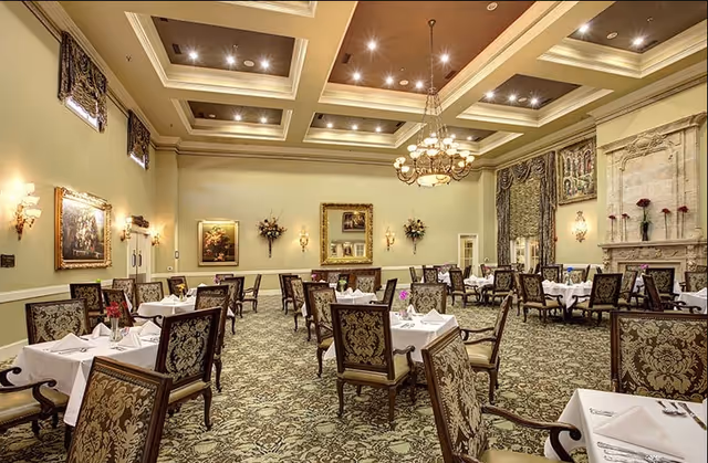 Elegant dining room with multiple tables covered in white tablecloths and set with napkins and silverware. The room features ornate patterned chairs, a decorative carpet, chandeliers hanging from a coffered ceiling, framed paintings on the walls, floral arrangements, and a large ornate fireplace.