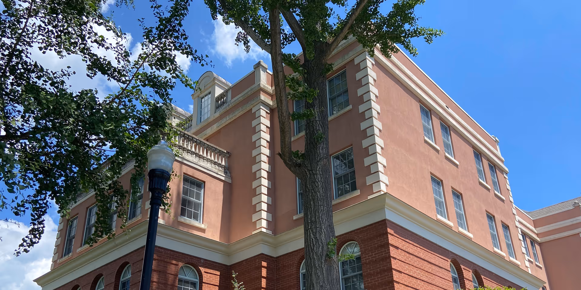 Exterior view of a multi-story senior living facility building with a combination of red brick and peach-colored walls, white trim, and multiple windows under a clear blue sky. A tree and a street lamp are visible in the foreground.