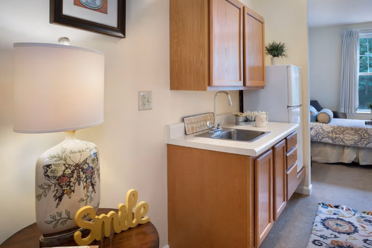 A small kitchenette area with wooden cabinets, a white countertop, and a stainless steel sink. To the left, there is a decorative table lamp with a butterfly design and a yellow 'Smile' sign on a wooden side table. In the background, a bedroom with a bed, pillows, and a window with curtains is visible. A colorful patterned rug is partially visible on the carpeted floor.