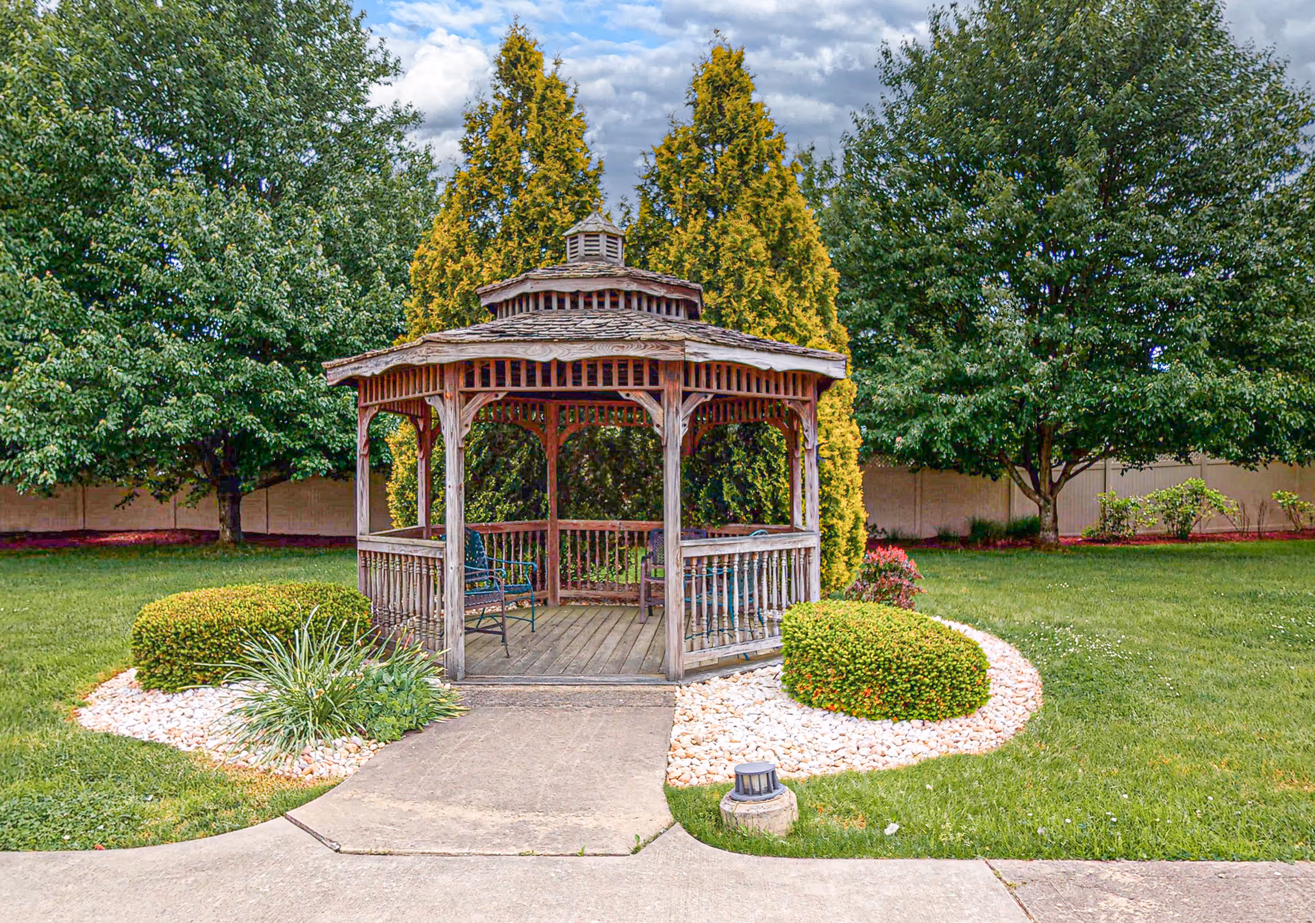 A wooden gazebo with a shingled roof sits in a well-maintained garden area. The gazebo has benches inside and is surrounded by neatly trimmed bushes, white decorative rocks, green grass, and several trees. The sky is partly cloudy.
