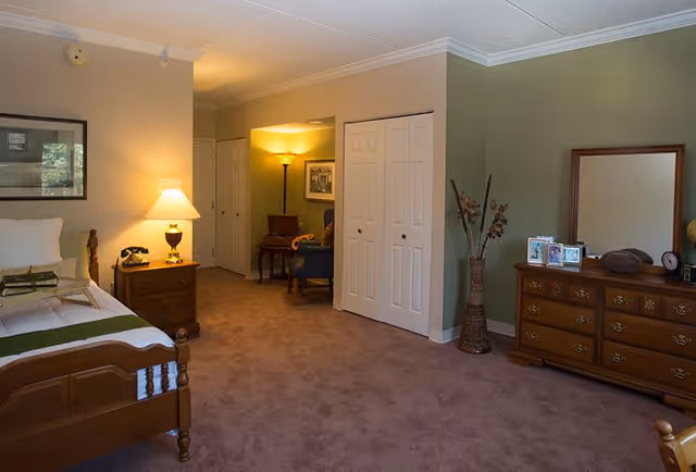 A cozy bedroom in a senior living facility featuring a wooden bed with white bedding and a green throw, a wooden nightstand with a lamp and a vintage telephone, a dresser with a mirror and framed photos, a tall vase with dried flowers, and a small sitting area with a chair and floor lamp in the background.