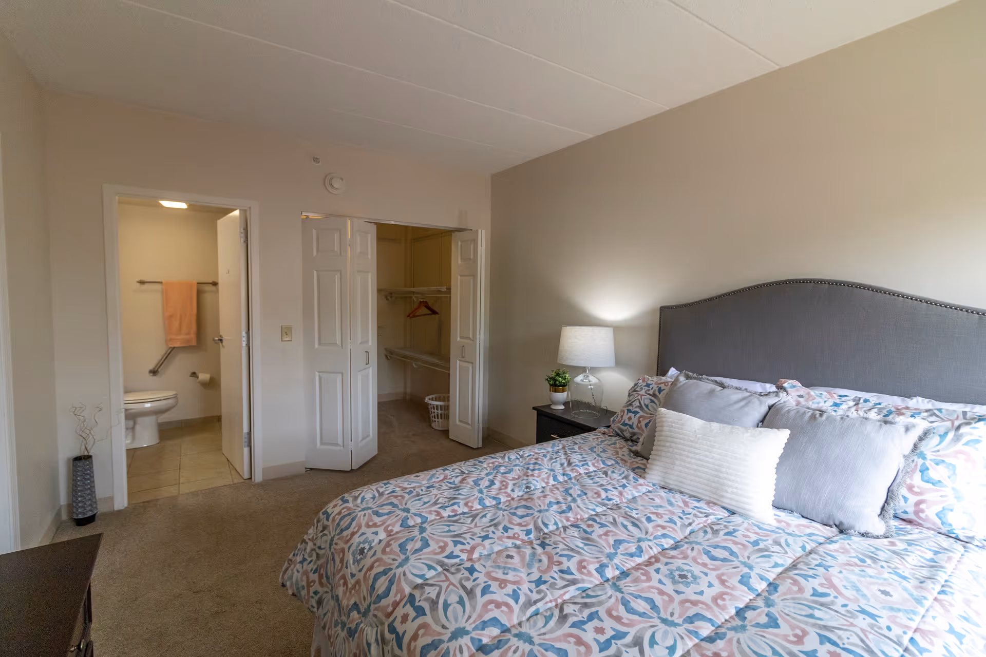 A bedroom with a patterned bedspread on a bed with multiple pillows, a nightstand with a lamp and a small plant, an open closet with shelves and hangers, and a bathroom with a toilet and a towel hanging on a rack.