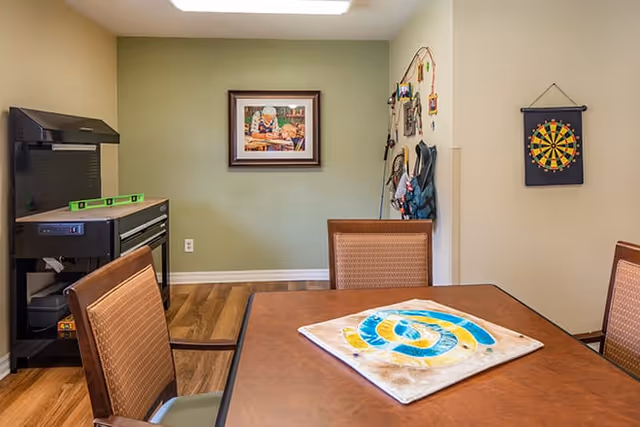A room with a wooden table and four chairs, a dartboard hanging on the wall, a framed picture on a green accent wall, and a black tool chest with a level on top. The floor is wood, and there are various items hanging on the wall near the corner.