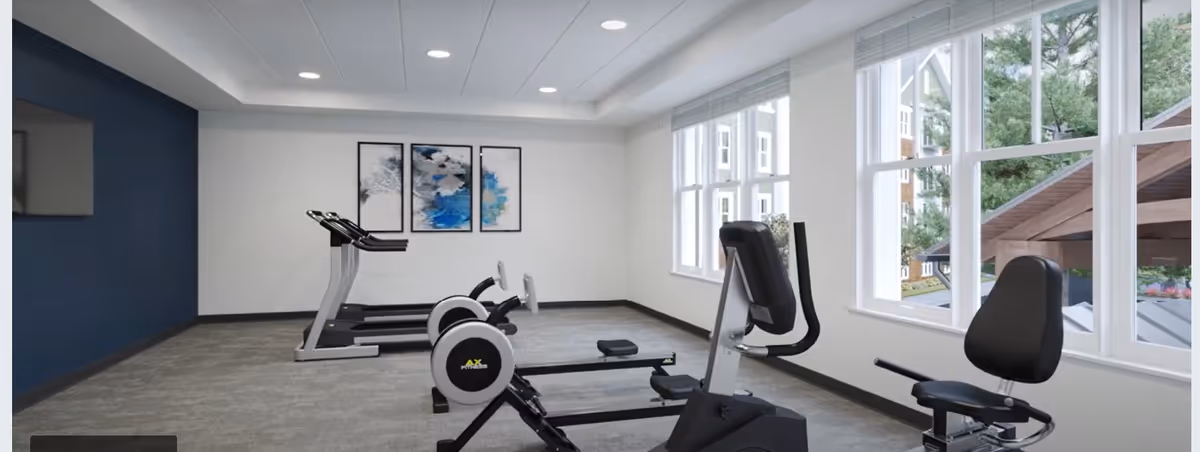 A bright fitness room with exercise equipment including a recumbent bike, rowing machines, and treadmills. The room has large windows letting in natural light, white walls, a blue accent wall, and three abstract paintings on the far wall.