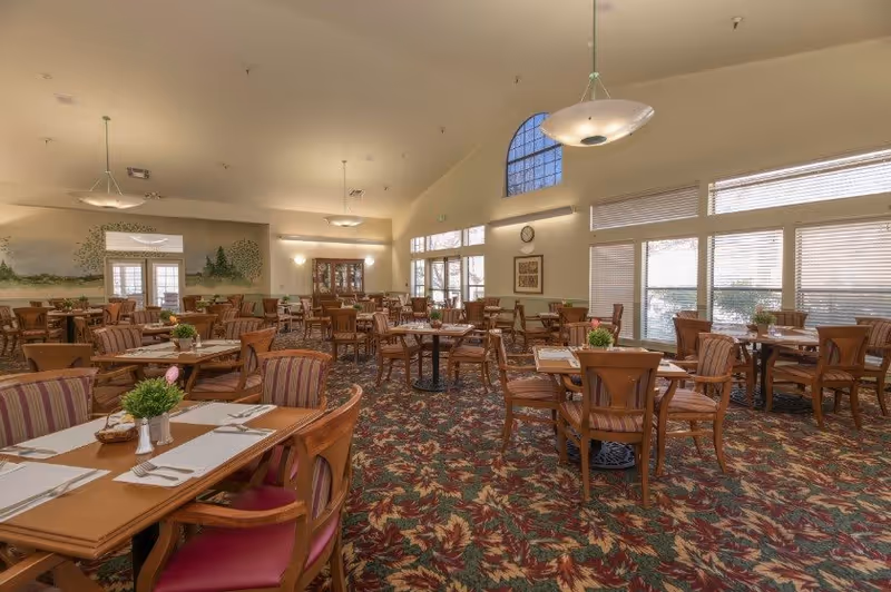A spacious senior living dining room with multiple wooden tables and chairs arranged neatly. Each table is set with placemats, napkins, and small green plants as centerpieces. The room features large windows with blinds, allowing natural light to fill the space. The ceiling is high with modern hanging light fixtures, and the carpet has a colorful leaf pattern. A mural of trees is visible on one wall, and a clock and framed artwork hang on another.