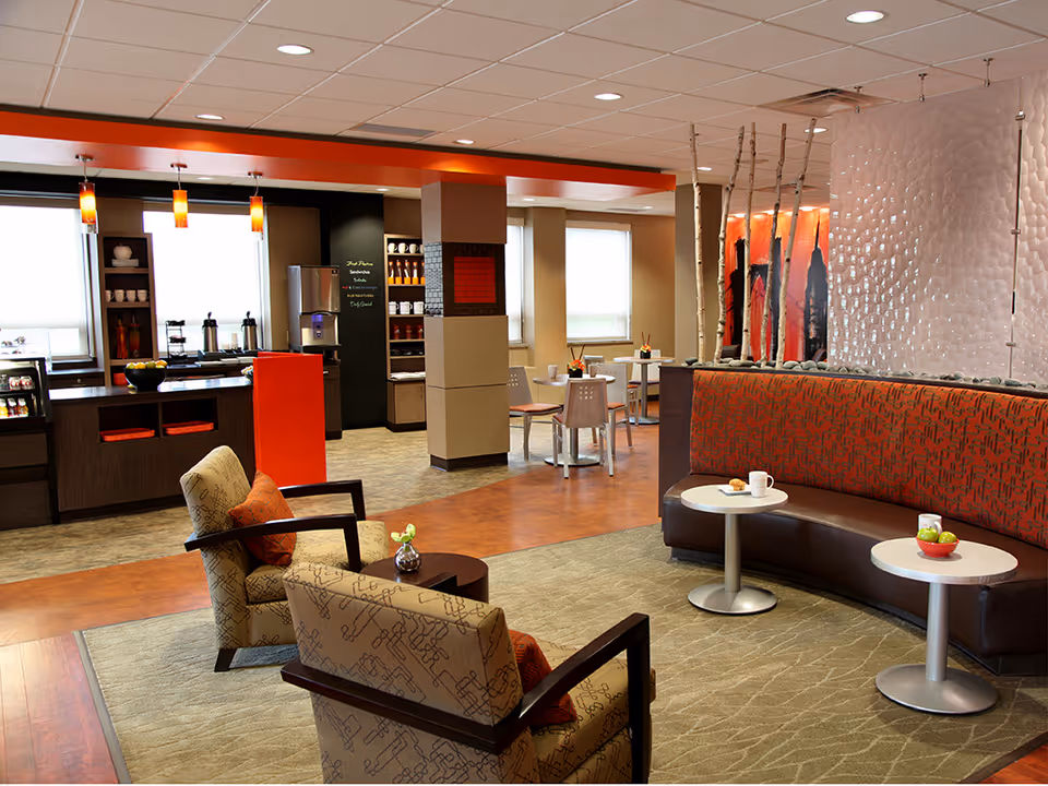 A modern and cozy common area in a senior living facility featuring comfortable armchairs with patterned upholstery and orange cushions, a curved bench with red and brown patterned upholstery, small round tables with cups and a bowl of fruit, and a kitchenette area with shelves, coffee dispensers, and pendant lights. The space has warm lighting, a mix of carpet and wood flooring, and decorative elements including tall sticks and a textured wall panel.