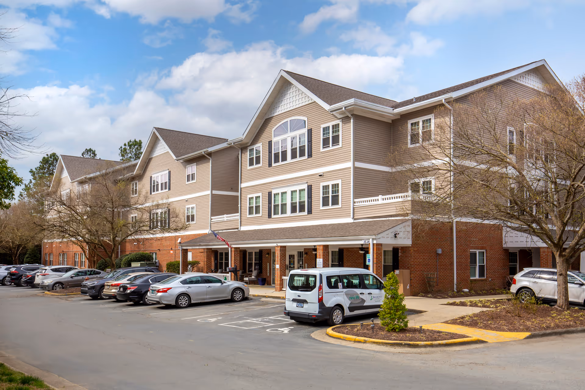Exterior view of a three-story senior living facility building with beige siding and brick accents. Several cars are parked in front of the building, including a white van with signage. The sky is partly cloudy and there are leafless trees around the parking area.