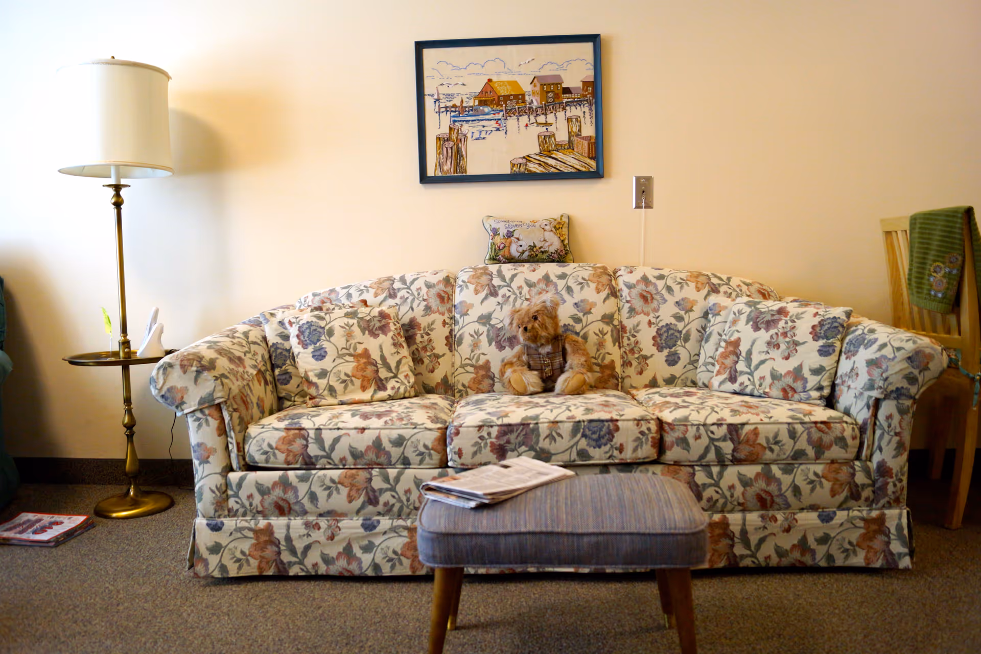 A floral patterned sofa with matching cushions and a teddy bear sitting in the middle. In front of the sofa is a small cushioned ottoman with a newspaper on top. To the left of the sofa is a tall brass floor lamp with a white lampshade and a small round side table. On the wall above the sofa hangs a framed painting of a dock scene. To the right, part of a wooden chair with a green cloth draped over it is visible.