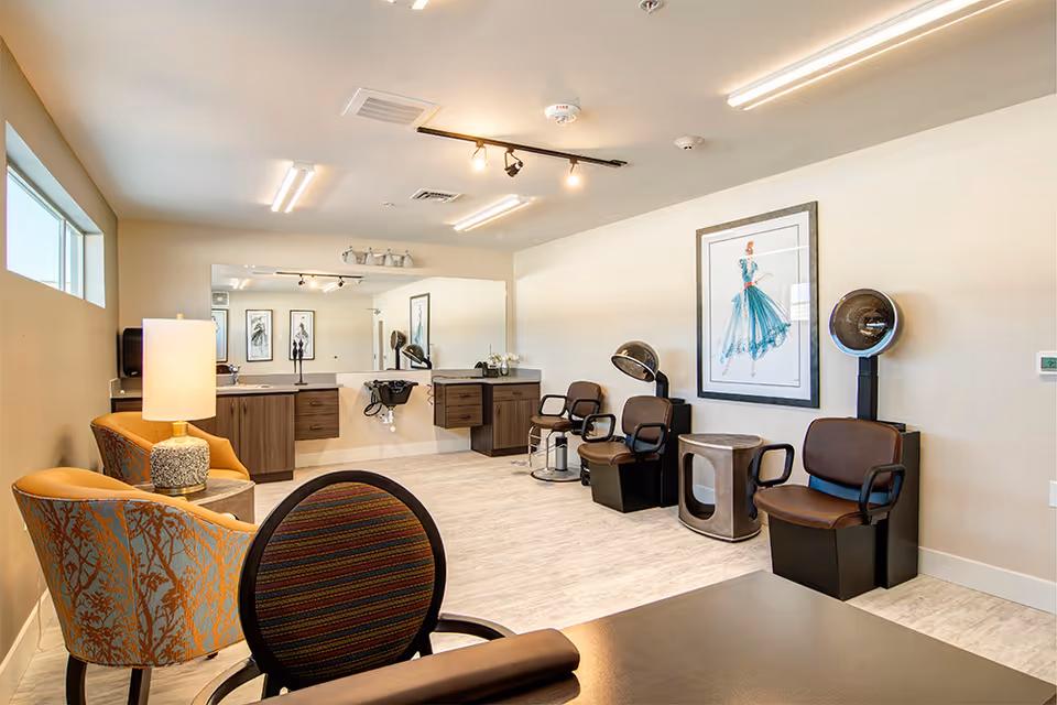 A bright and clean salon area in a senior living community featuring three brown salon chairs with hair dryers, a large mirror above a counter with a sink, two patterned armchairs with a lamp on a small table between them, and a framed artwork of a woman in a blue dress on the wall.