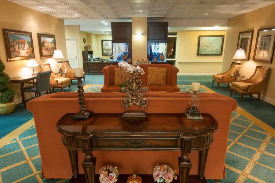 A cozy senior living facility common area with orange sofas, a wooden console table adorned with decorative items and candles, upholstered chairs, framed paintings on the walls, and soft lighting from table lamps. The space has a patterned carpet and a welcoming atmosphere.