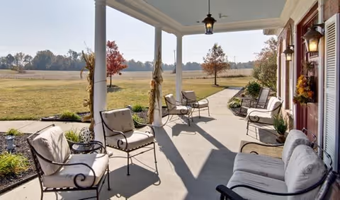 A covered outdoor patio area with cushioned metal chairs and small tables, overlooking a grassy field with a few trees in the distance under a clear sky.