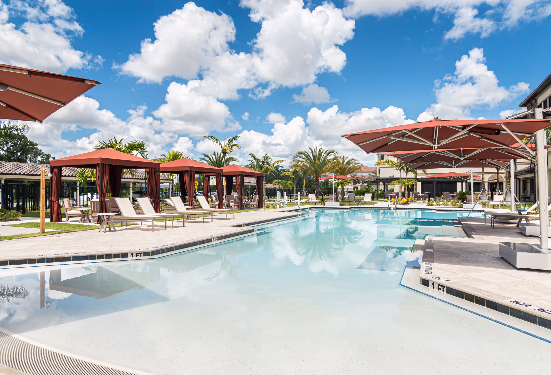 Outdoor swimming pool area with several lounge chairs and cabanas with red canopies. Palm trees and other greenery surround the pool under a bright blue sky with scattered white clouds.