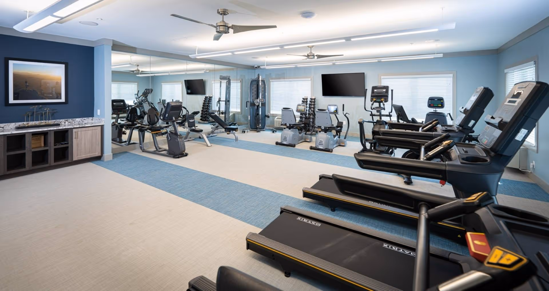 A spacious fitness room with several treadmills, exercise bikes, weight machines, and free weights. The room has light blue walls, large windows with blinds, ceiling fans, and a wall-mounted TV. There is a mirrored wall reflecting the equipment and a countertop with cabinets on the left side.