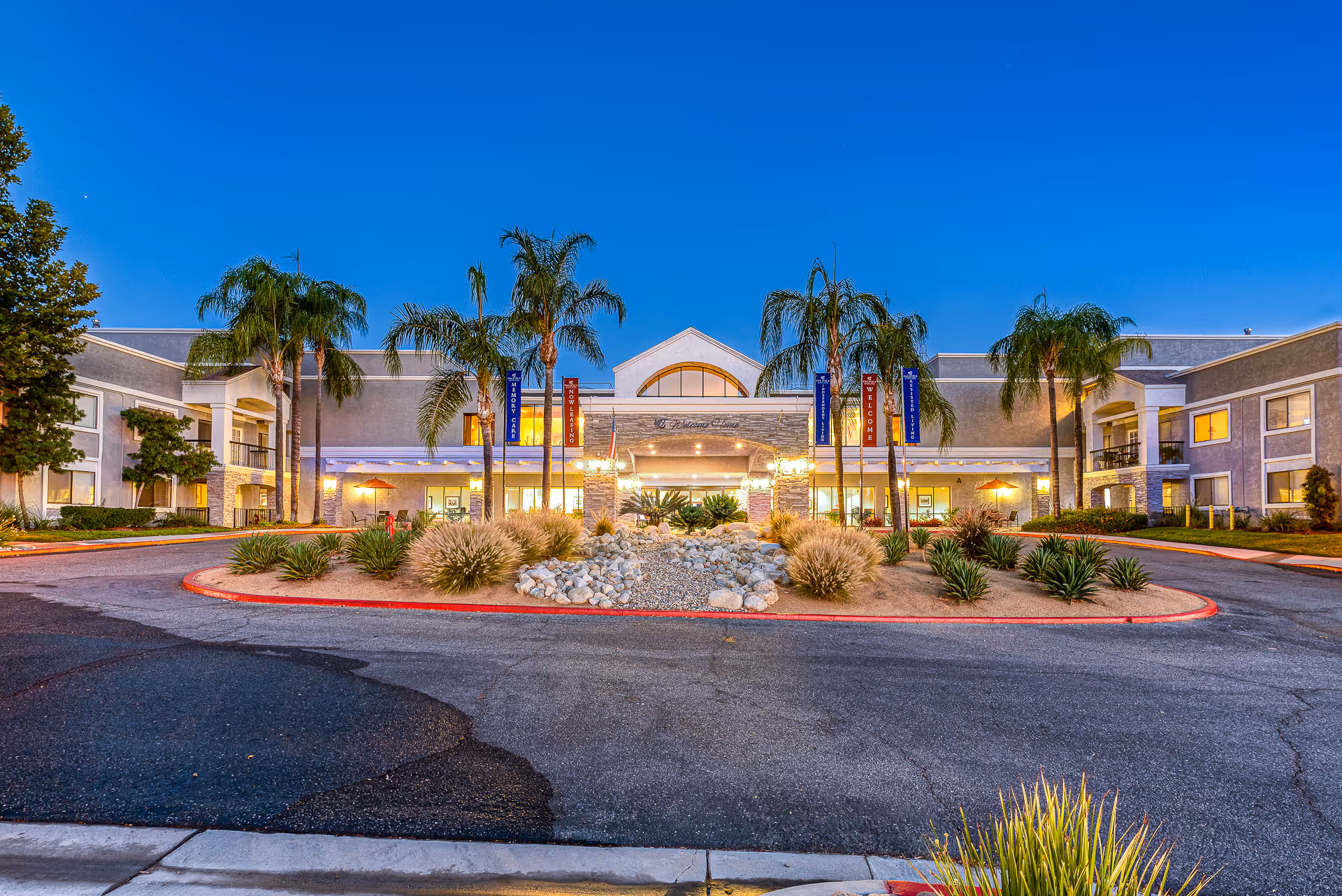 Front exterior view of Menifee Senior Living facility at dusk with palm trees, landscaped roundabout with rocks and plants, and building lights on.