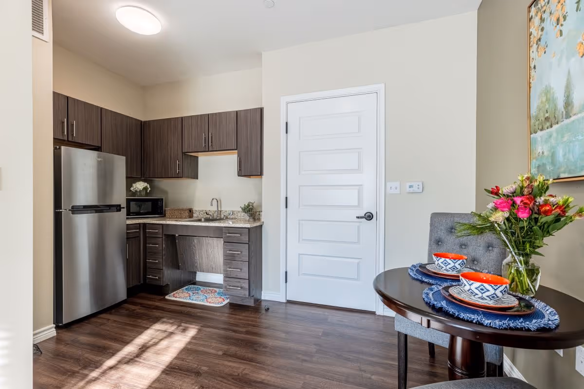 A small kitchen area with dark wood cabinets, a stainless steel refrigerator, a microwave, and a sink with a granite countertop. To the right, there is a round dining table set for two with colorful bowls, plates, and a vase of flowers. The floor is dark wood, and a white door is visible in the background.