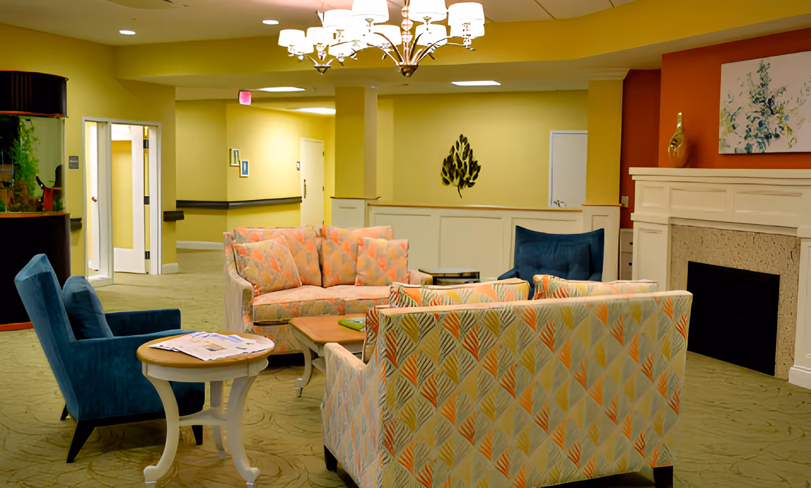 A cozy living room area in a senior living facility with patterned sofas, two blue armchairs, a wooden coffee table, a side table with newspapers, a fireplace, and wall decorations including a painting and a metal leaf sculpture. The walls are painted yellow and orange, and there is a chandelier hanging from the ceiling.
