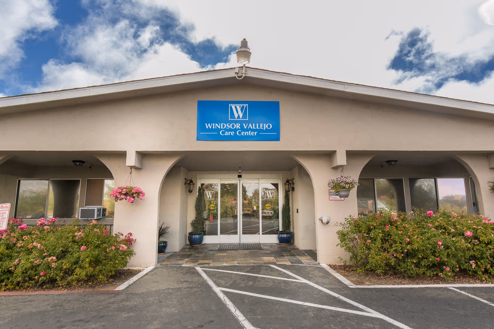 Front exterior view of Windsor Vallejo Care Center building with a beige facade, a blue sign above the entrance, glass double doors, hanging flower pots, and bushes with pink flowers on either side of the entrance.