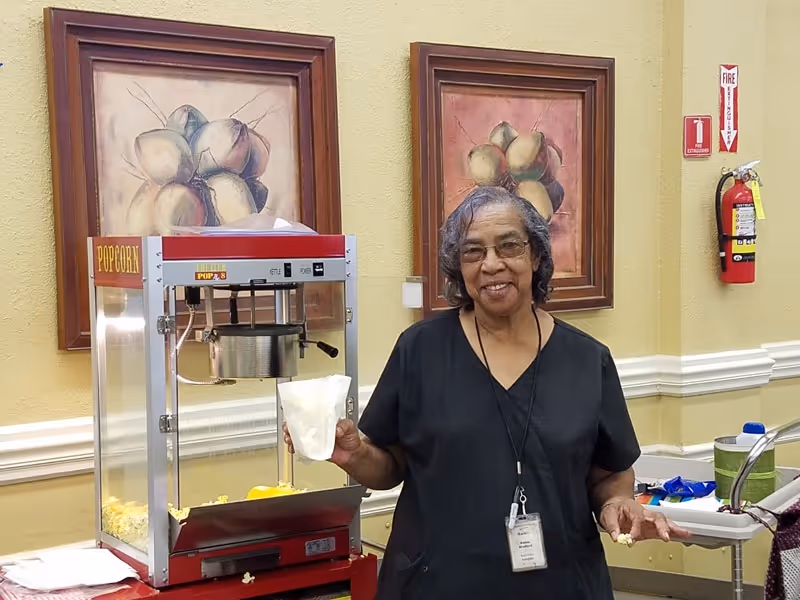 An elderly woman wearing glasses and a black shirt stands next to a popcorn machine, holding a bag of popcorn in one hand and some popcorn in the other. Behind her, there are two framed paintings of fruit on a beige wall, and a fire extinguisher is mounted on the wall to the right.