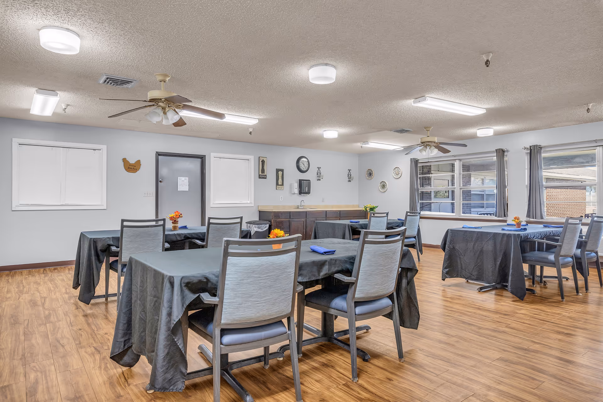 A dining room in a senior living facility with several tables covered in black tablecloths, each set with a blue napkin and small flower arrangements. The room has wooden flooring, ceiling fans, fluorescent lighting, and large windows with gray curtains letting in natural light. The walls are decorated with clocks and small framed pictures.