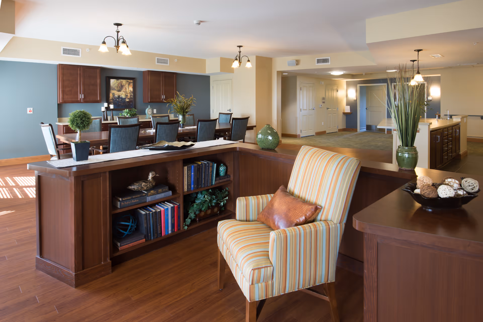 A spacious and well-lit common area with wooden flooring featuring a striped armchair with a brown pillow, a wooden bookshelf with books and decorative items, a long dining table with chairs, and decorative plants. The walls are painted in soft beige and blue tones, and ceiling lights illuminate the room.