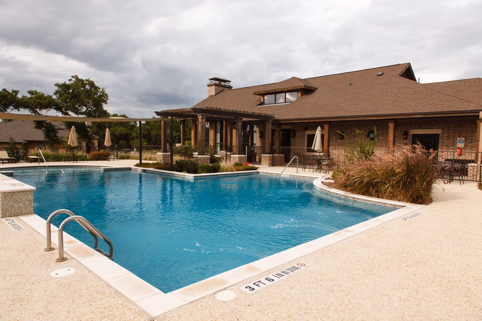 Outdoor swimming pool with clear blue water surrounded by a beige concrete deck. There are metal handrails for pool entry, several patio tables with umbrellas, and a brick building with a brown roof in the background under a cloudy sky.