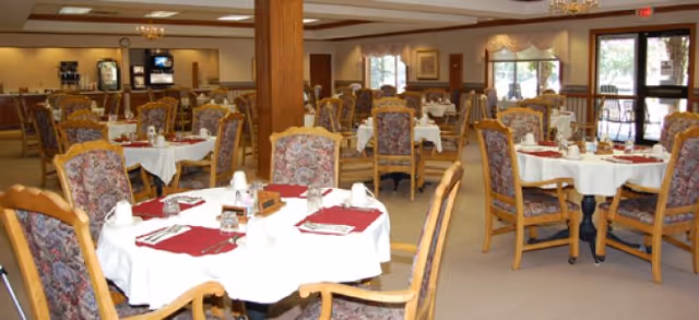 A spacious dining room with multiple round tables covered in white tablecloths and red placemats, each set with napkins, glasses, and silverware. The room features cushioned wooden chairs with floral upholstery, large windows letting in natural light, and a beverage station in the background.