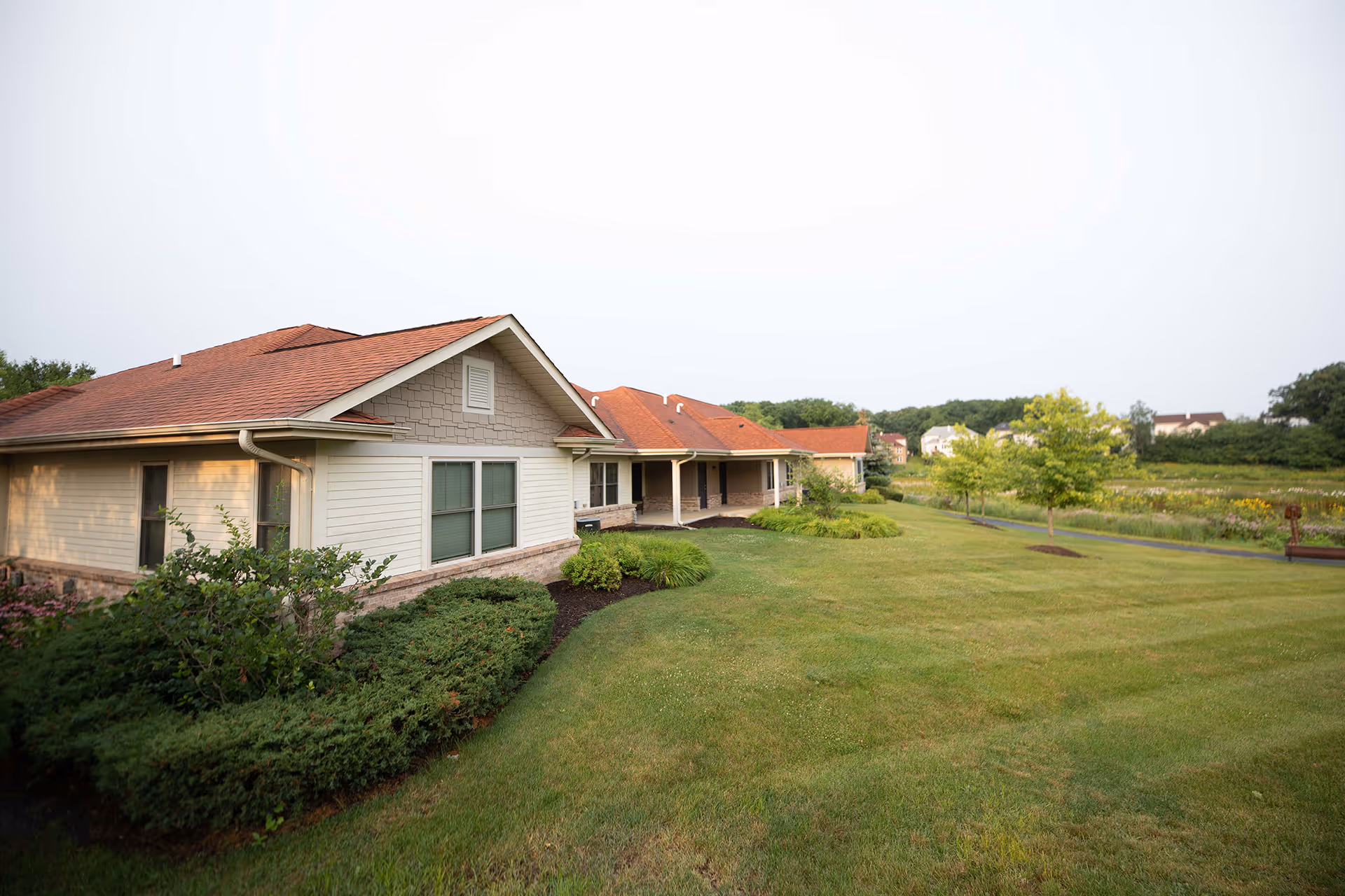 Exterior view of a single-story building with a red roof and beige siding, surrounded by well-maintained green lawns, shrubs, and trees under a clear sky.