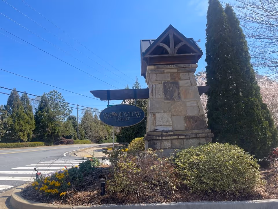 Stone entrance sign reading "Woodview Court" beside landscaped shrubbery and a roadway under a clear blue sky.