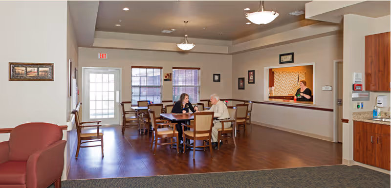 A spacious common area in a senior living facility with wooden flooring and beige walls. There are several wooden tables and chairs arranged in the room. Two people are seated at one of the tables engaged in conversation. A woman stands behind a counter window, possibly serving or assisting. The room is well-lit with ceiling lights and natural light coming through two large windows. A red armchair and additional chairs are visible along the walls.